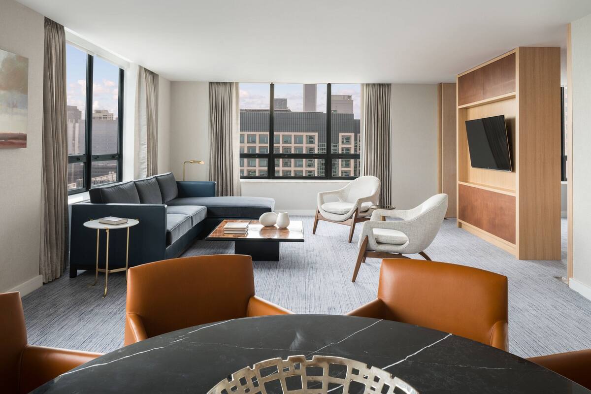 A modern hotel room with a sofa, chairs, a dining table, and a TV.