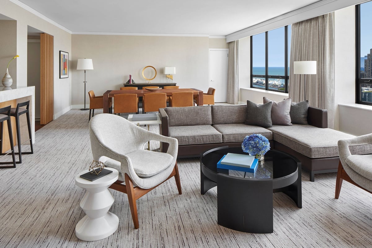 A modern hotel room with a living area, dining space, and a large window with a city view.