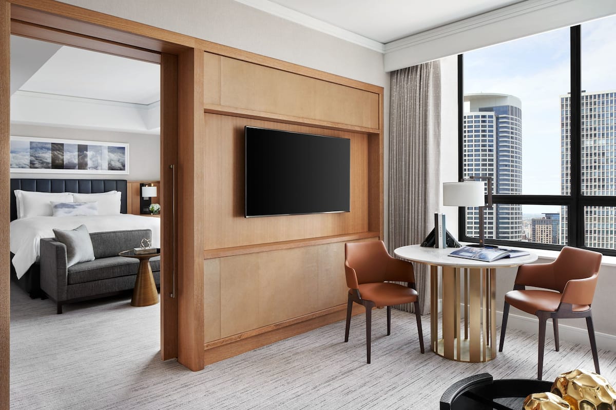 A hotel room with a bed, a TV mounted on a wooden panel, a round table with chairs, and a view of city buildings through large windows.