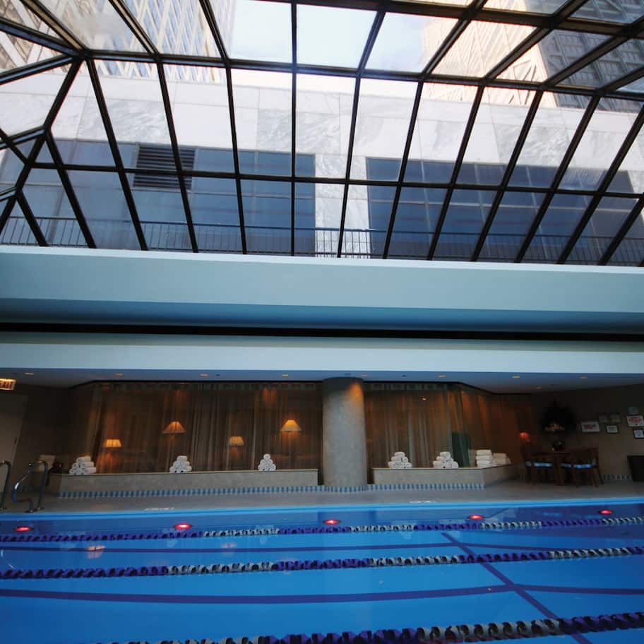 A swimming pool with a glass ceiling, stone statues, and a seating area with tables and chairs.