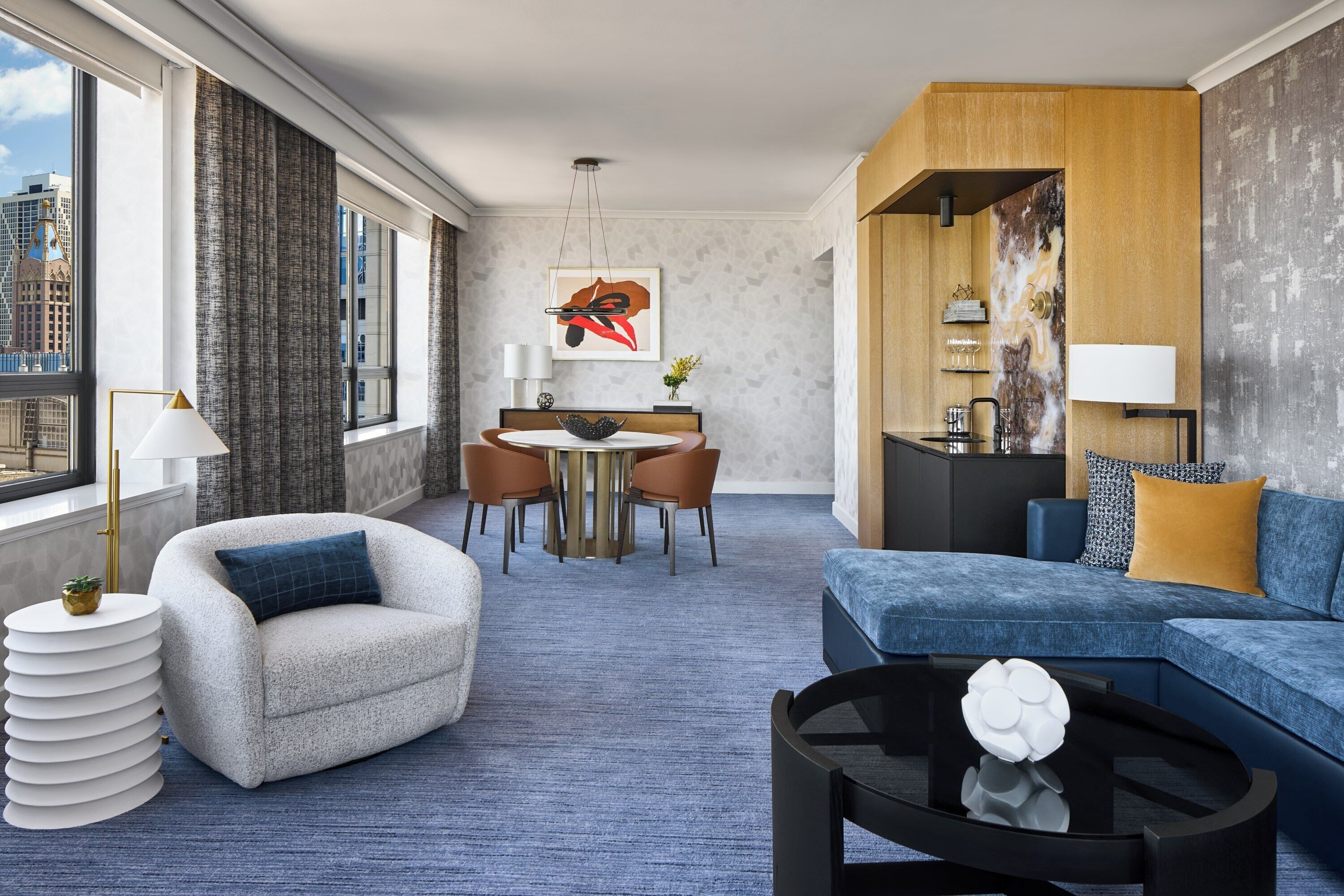 A modern hotel room with a dining area, a sofa, a coffee table, and a view of the city.
