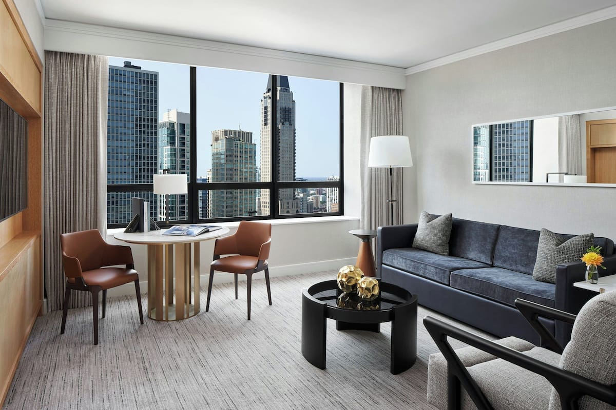 A modern hotel room with a large window offering a city view, a dark blue sofa, a round coffee table with decorative items, and a small side table with a lamp.