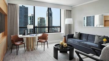 A modern hotel room with a large window offering a city view, a dark blue sofa, a round coffee table with decorative items, and a small side table with a lamp.