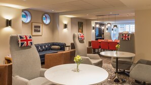Un hall d’hôtel avec un canapé bleu, des fauteuils gris, une table ronde ornée d’un vase et un coin repas avec des chaises rouges.