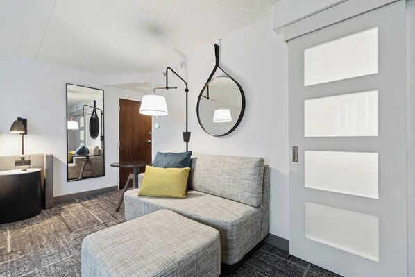 Habitación de hotel moderna con sofá gris, reposapiés y un espejo redondo. Cuenta con un escritorio con lámpara y una mesita redonda. La habitación tiene una puerta de madera y un amplio ventanal con persianas.