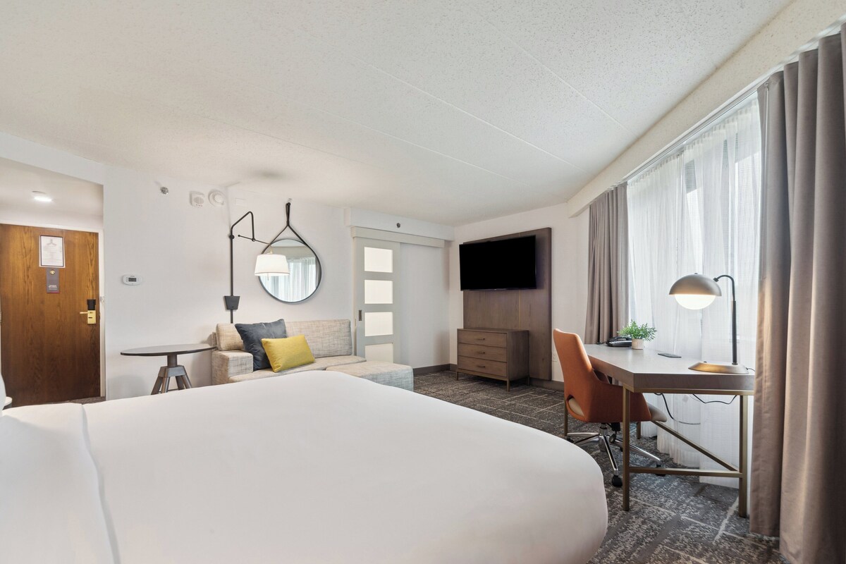 A hotel room with a large bed, a desk with a chair, a TV, and a window with curtains.