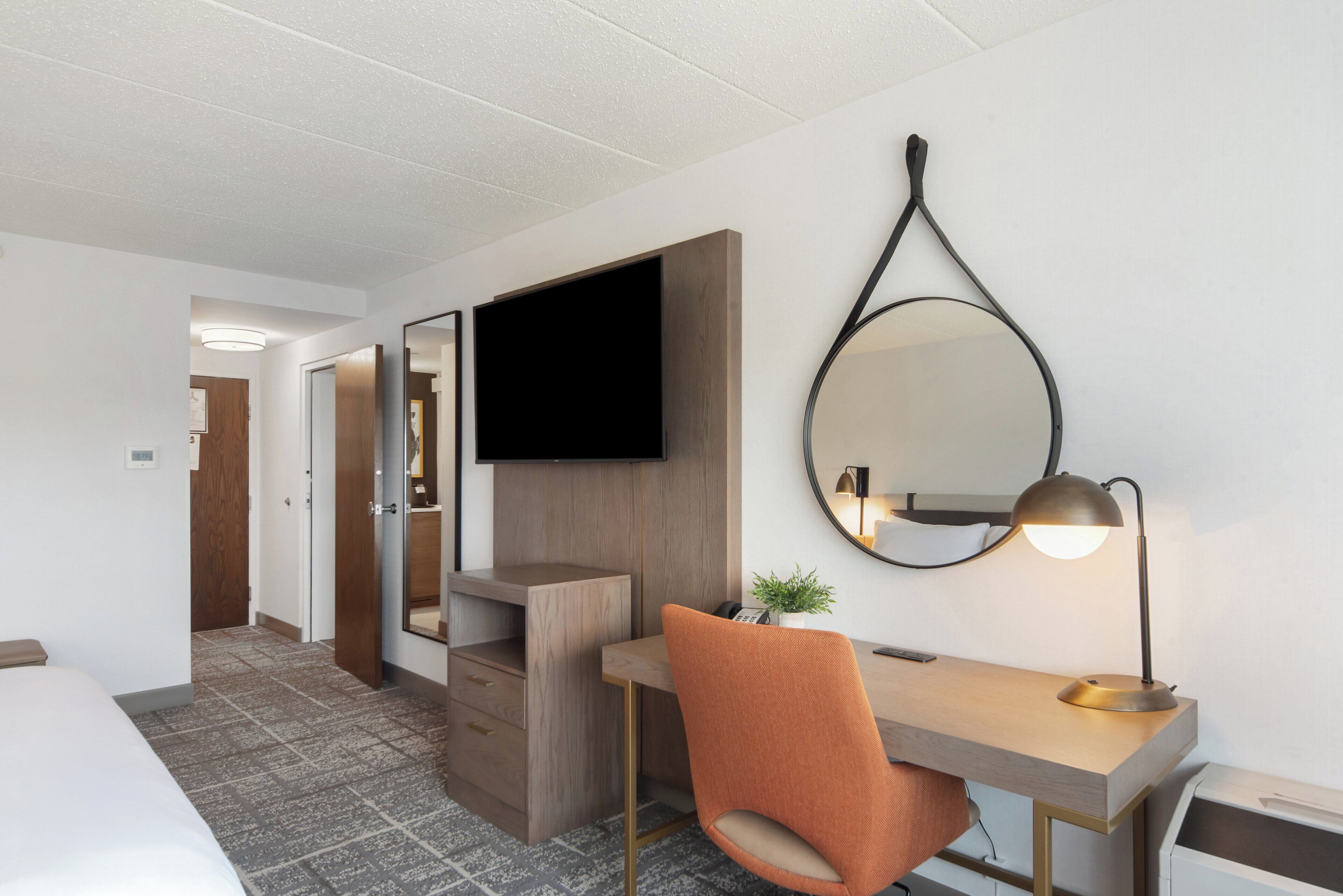 Quarto de hotel com cama, escrivaninha com cadeira, televisão de tela plana, espelho e abajur.