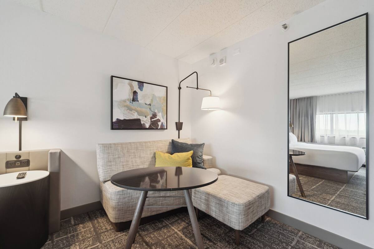 A modern hotel room with a sofa, a small round table, a lamp, a painting, and a large mirror reflecting a bed and window.