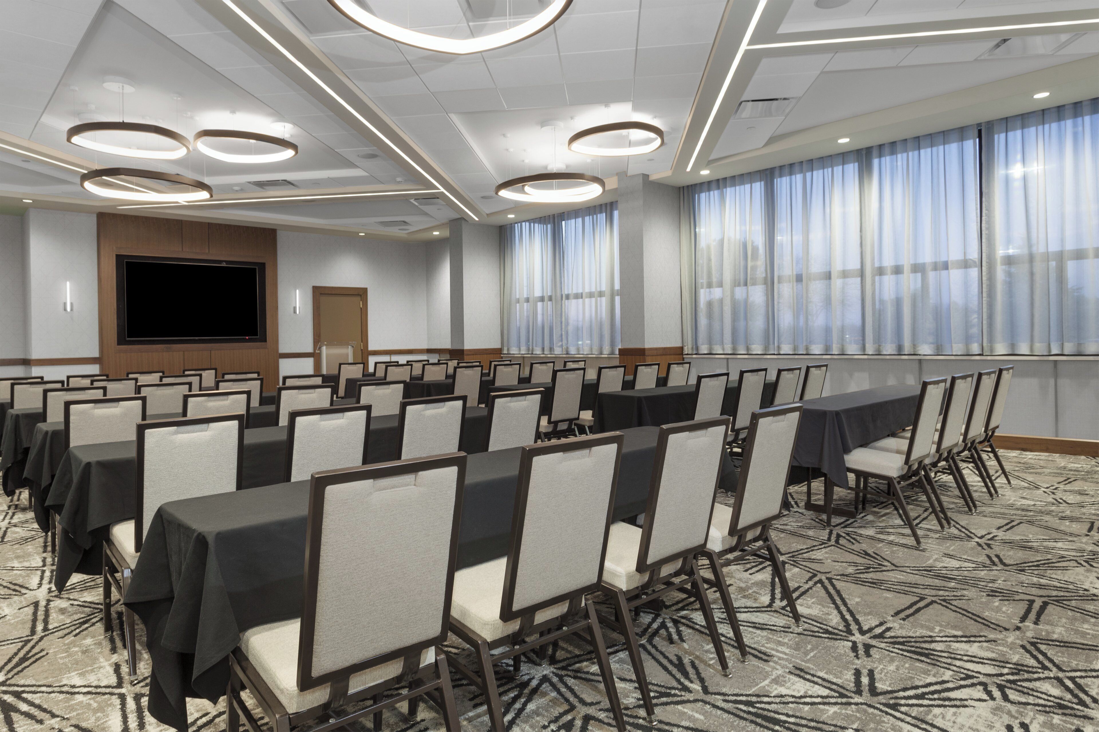A conference room with rows of tables and chairs, a large screen, and a window with curtains.