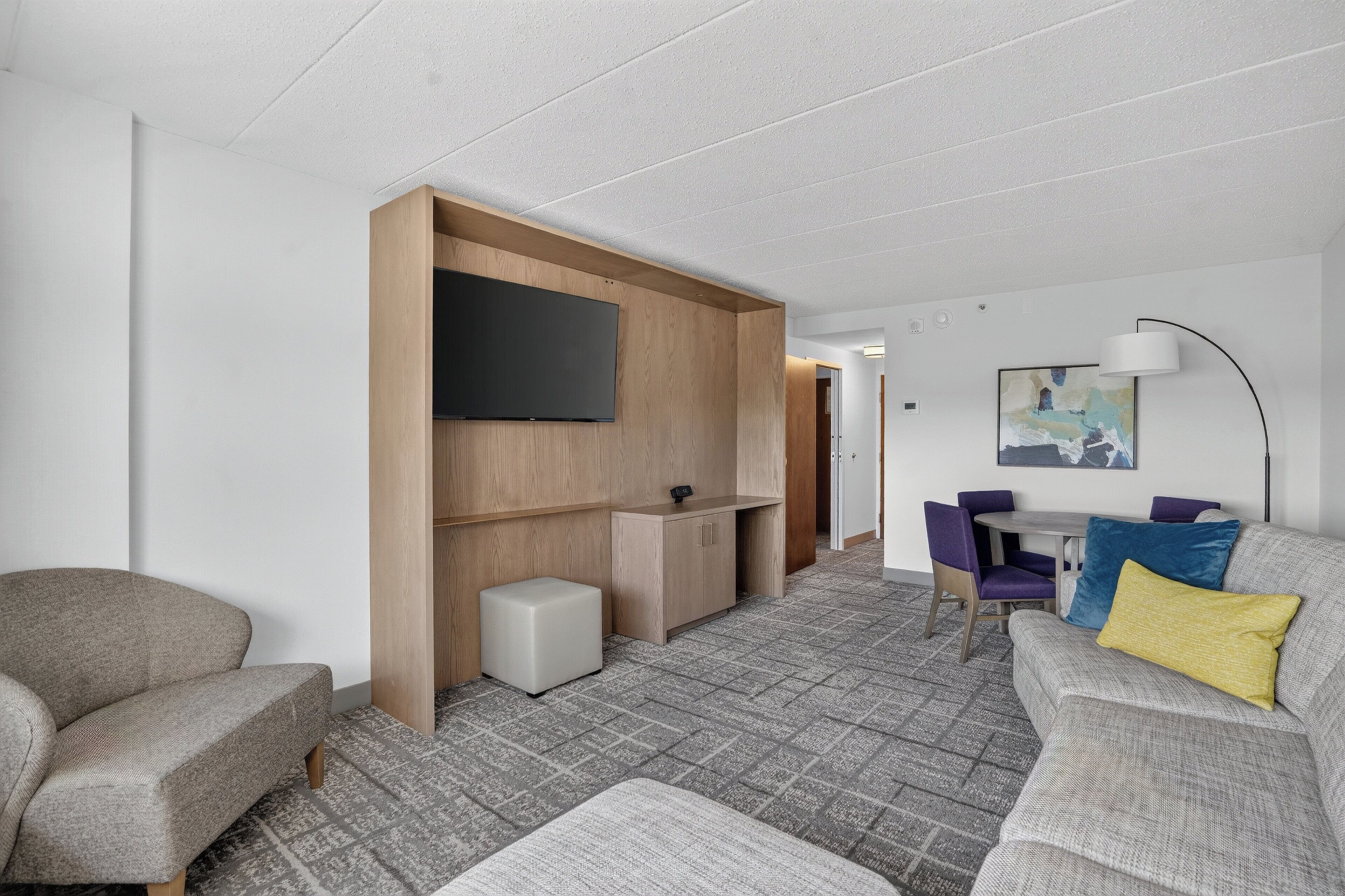 A modern hotel room with a sofa, a small table, a flat-screen TV mounted on a wooden partition, and a dining area with chairs.