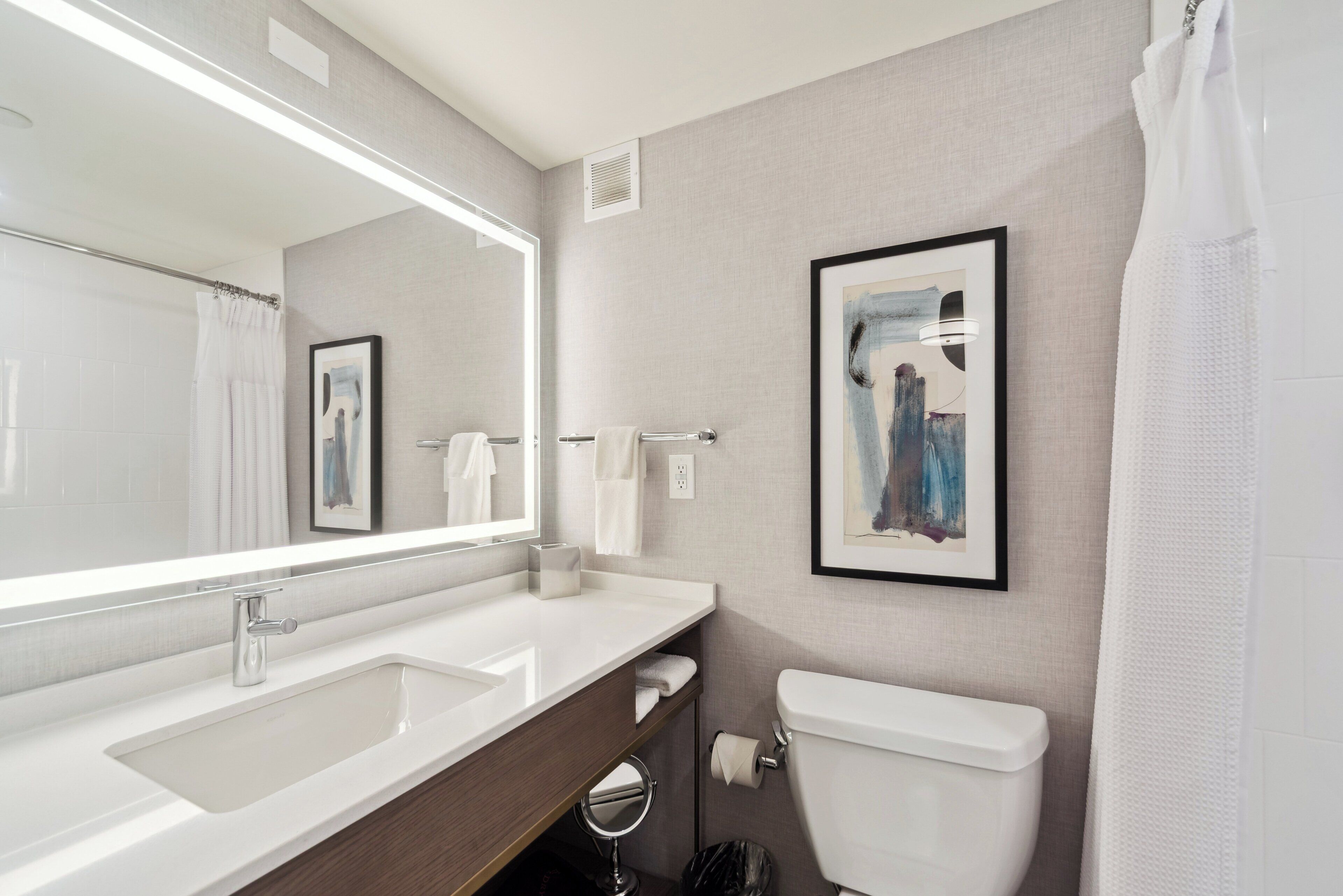 A modern bathroom with a large mirror, two sinks, a toilet, and a framed abstract painting on the wall.