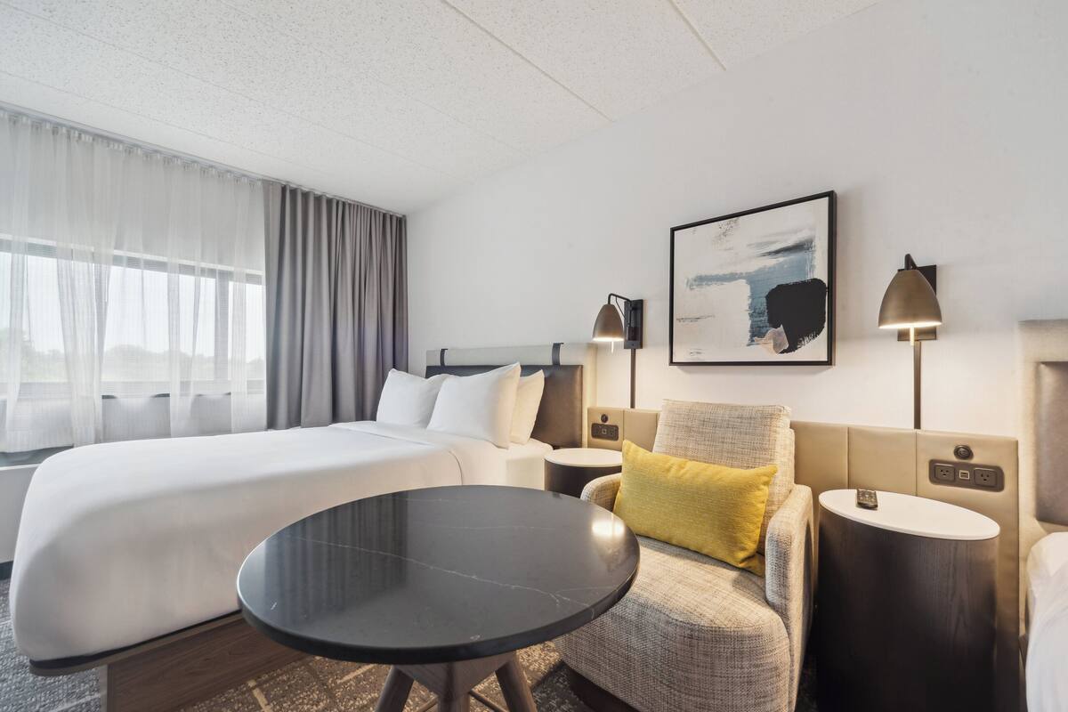 A hotel room with a large bed, a round table, a sofa with a yellow pillow, two bedside tables with lamps, and a framed abstract painting on the wall.