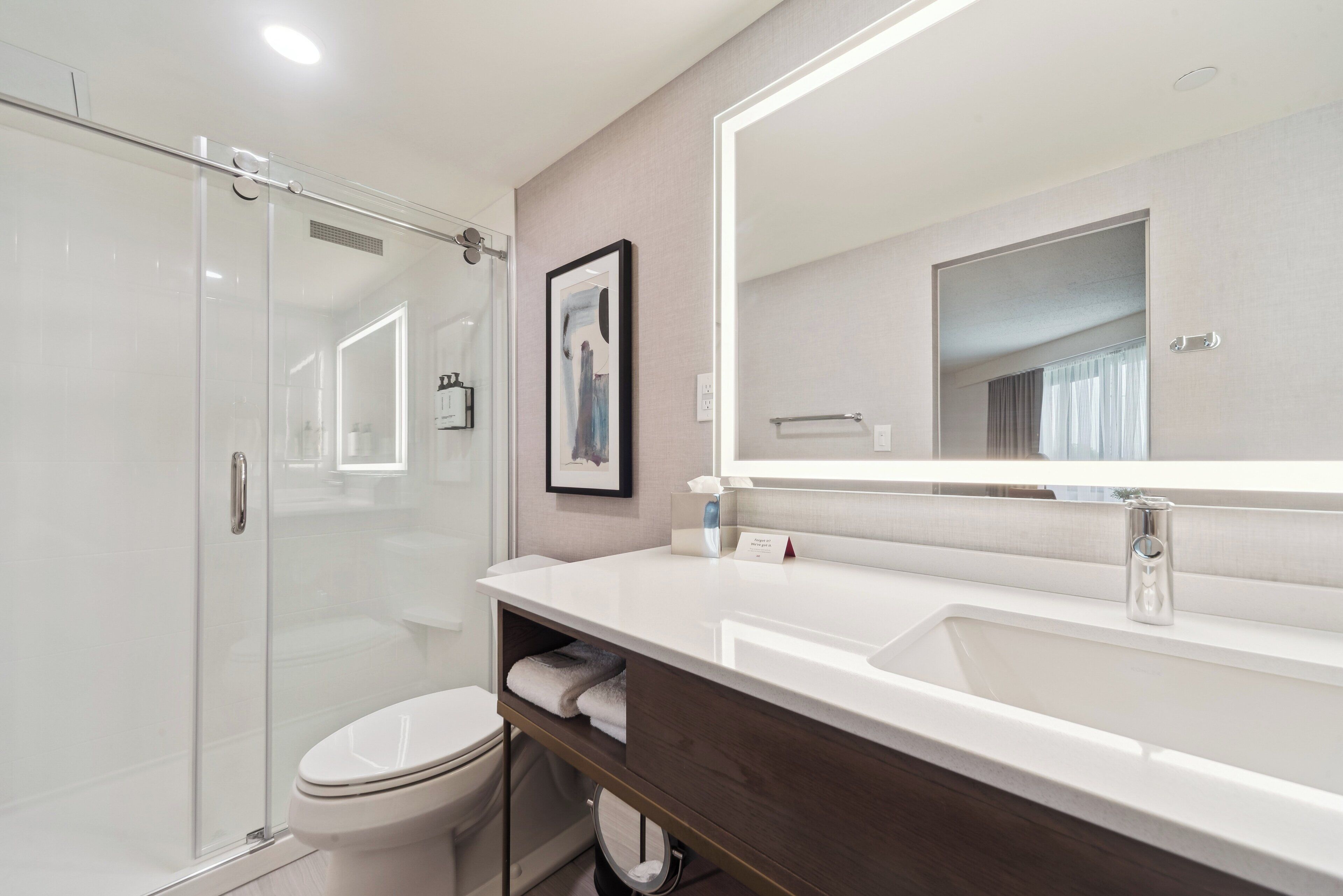 Un baño moderno con cabina de ducha de vidrio, inodoro blanco, tocador con un espejo grande y un perchero para toallas.
