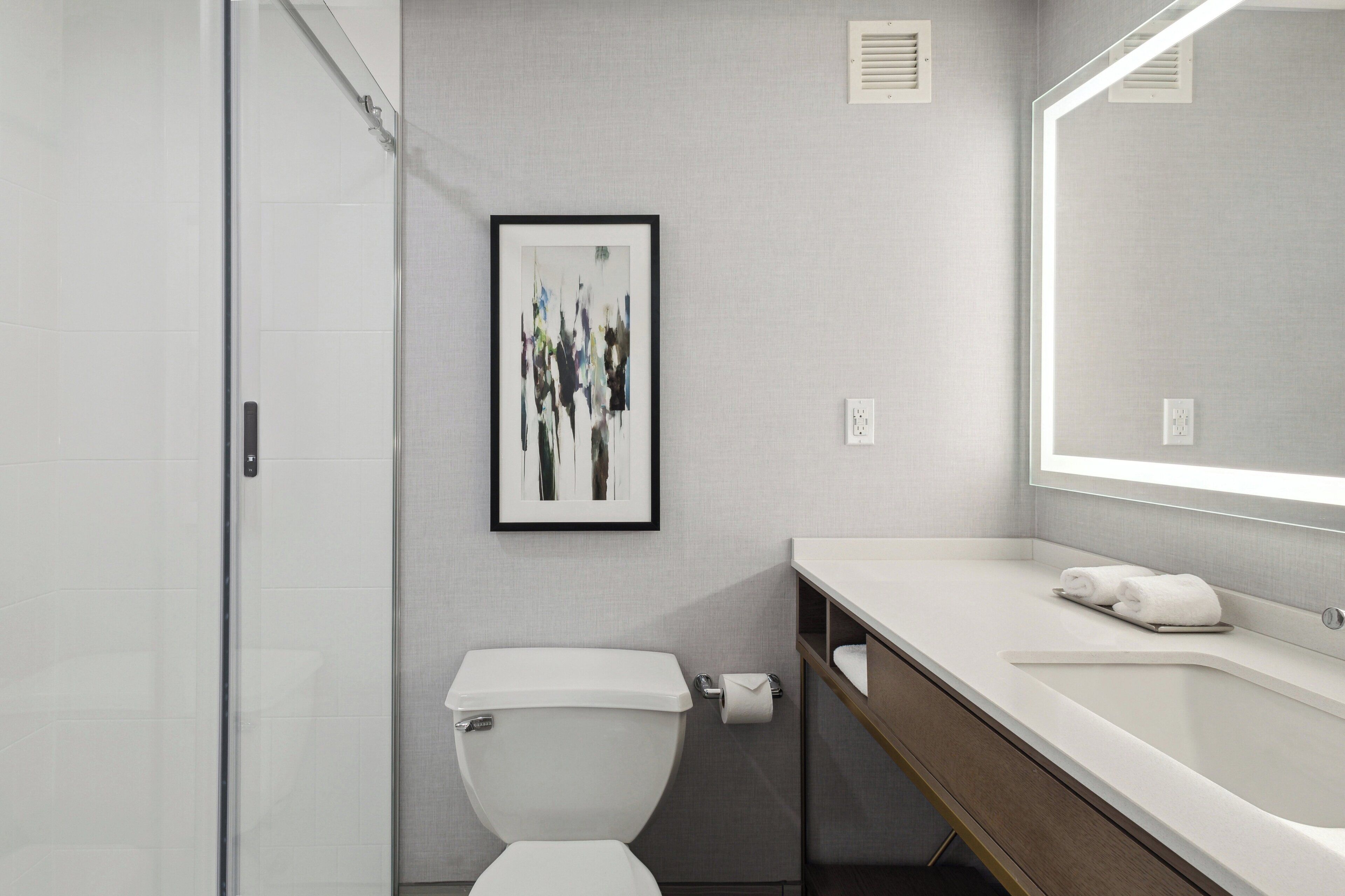 A modern bathroom with a glass shower enclosure, a toilet with a white lid, a framed abstract artwork on the wall, and a large mirror above the sink.