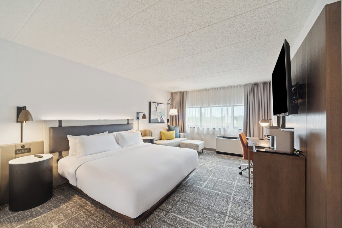 A hotel room with a large bed, a desk, a TV, and a window with curtains.