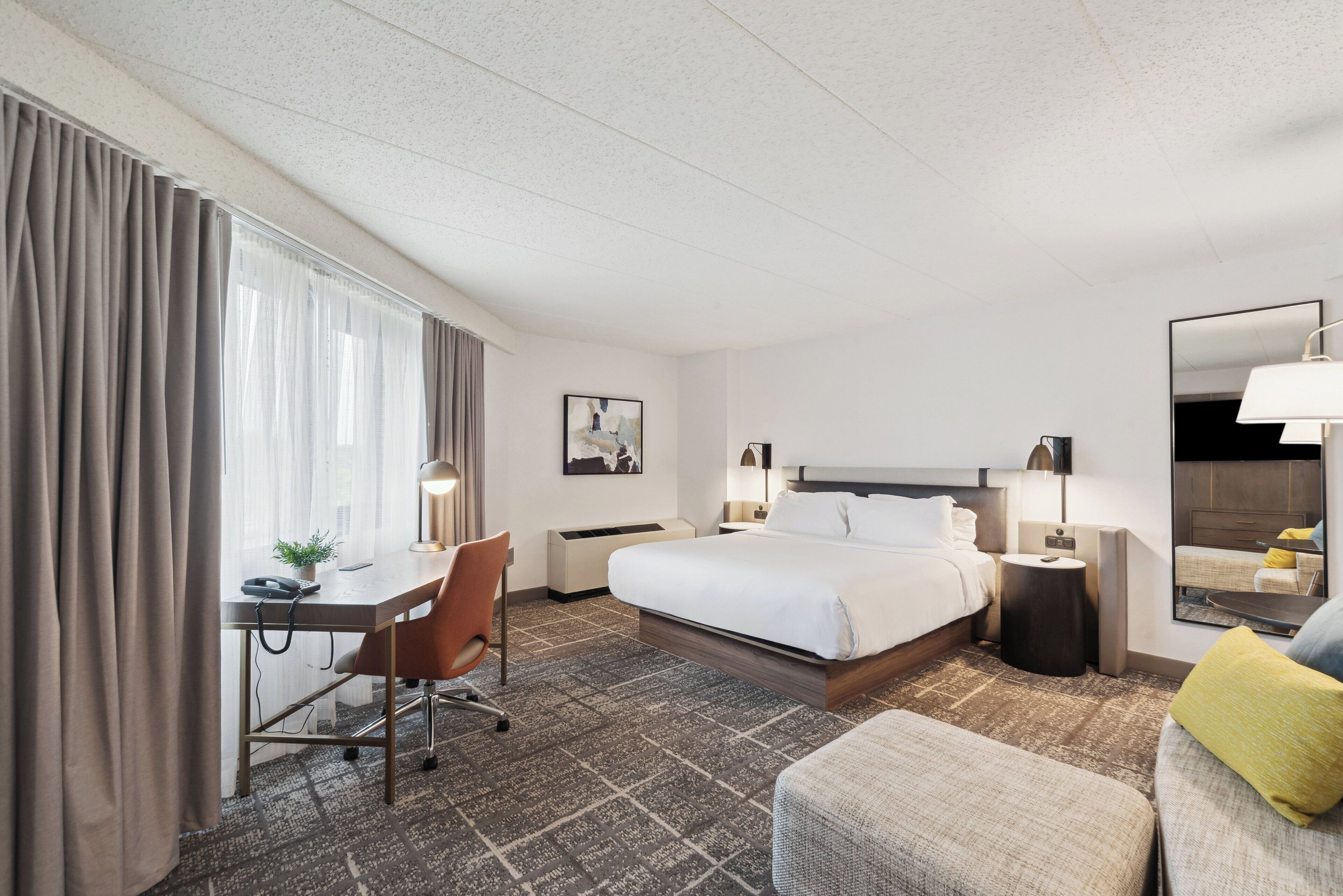 Une chambre d’hôtel avec un grand lit, un bureau avec une chaise, une petite table, une lampe de chevet, un miroir et une fenêtre avec des rideaux.