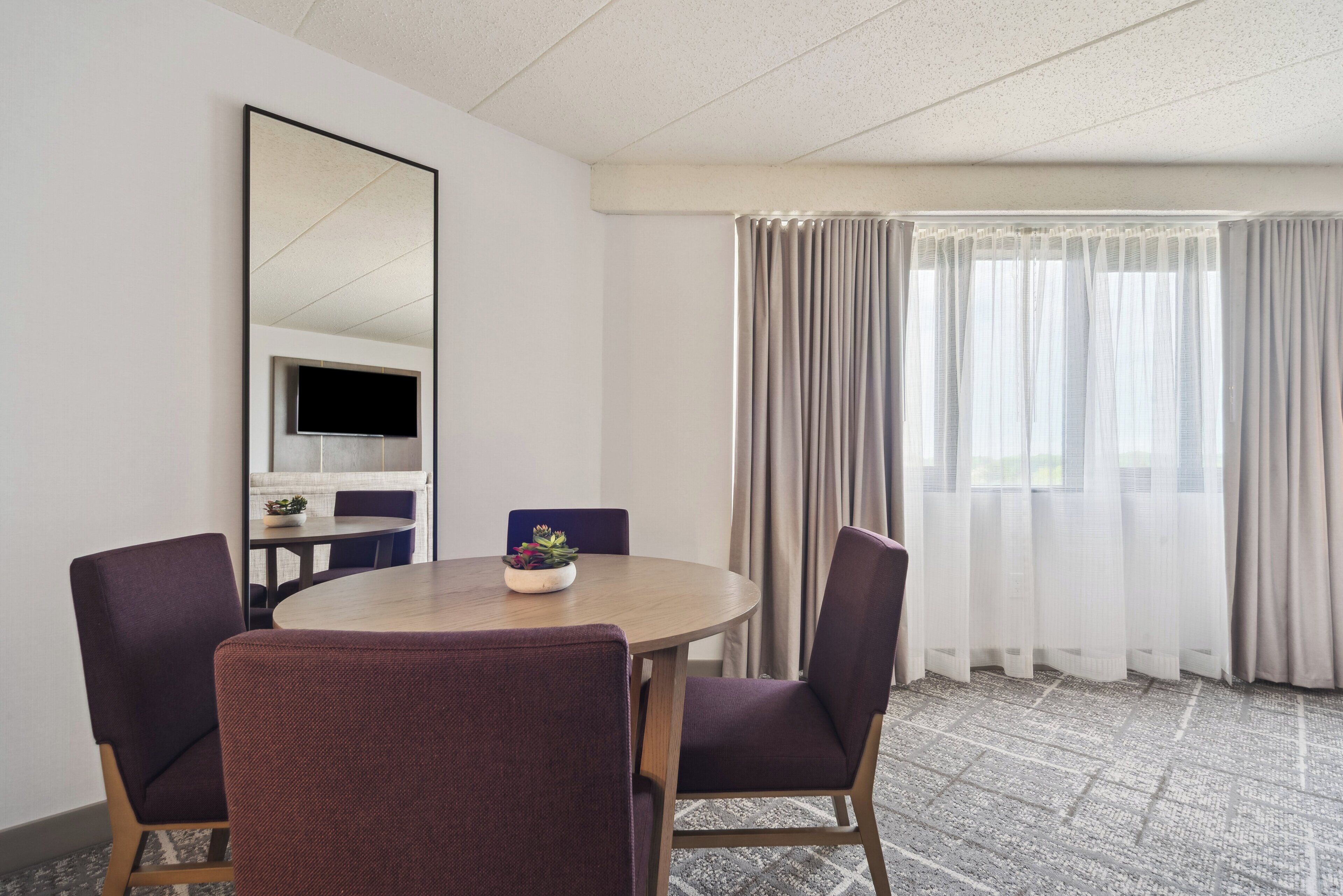A conference room with a round table, four chairs, a TV, and a window with curtains.