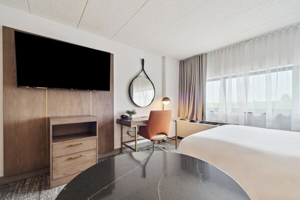 A hotel room with a large bed, a desk with a chair, a flat-screen TV mounted on the wall, a mirror, and a window with curtains. - Crowne Plaza Lansing by IHG (Lansing)
