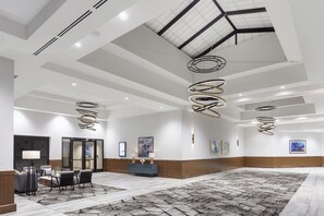 A spacious hotel lobby with a high ceiling, modern lighting fixtures, and a large seating area. - Crowne Plaza Lansing by IHG (Lansing)