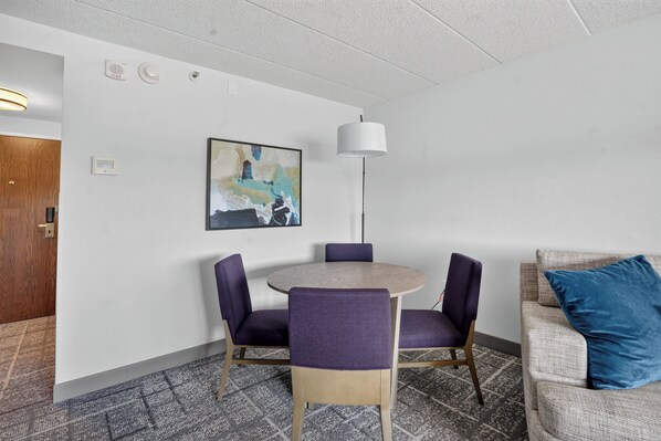 Une chambre d’hôtel avec une table ronde, des chaises violettes, un lampadaire blanc, un tableau au mur et un coussin bleu sur un canapé.