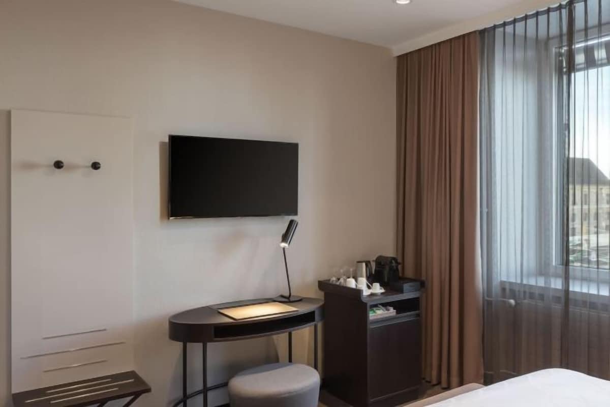 A hotel room with a bed, a desk with a chair, a TV mounted on the wall, and a window with curtains.