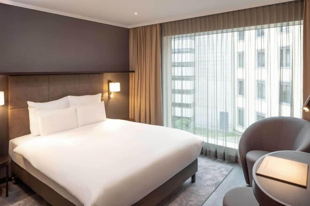 A hotel room with a large bed, a bedside lamp, a chair, and a view of buildings through the window.