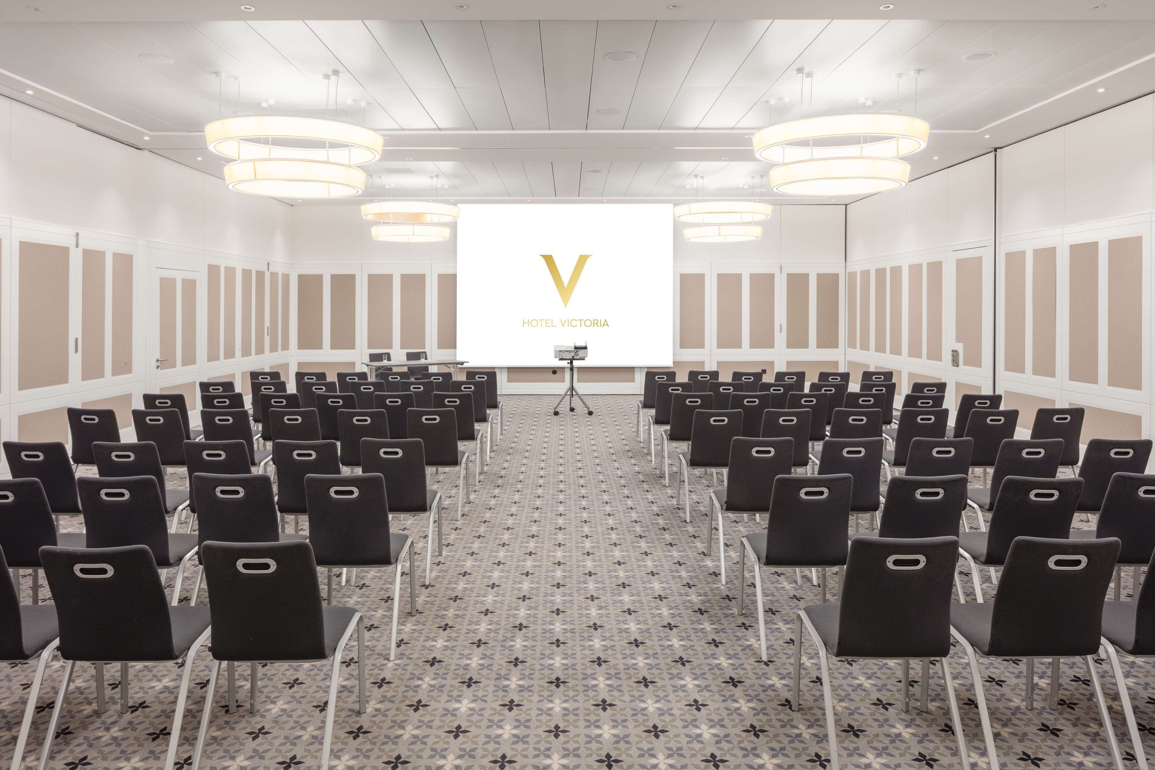 A conference room with rows of chairs, a podium, and a large screen displaying the logo of Hotel Victoria.