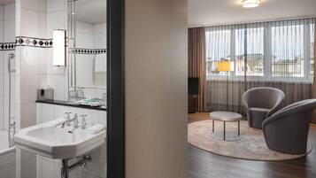 Ein modernes Badezimmer mit Waschbecken, Spiegel und Dusche. Angrenzend befindet sich ein Wohnbereich mit einem runden Tisch, zwei Stühlen und Blick durch große Fenster.