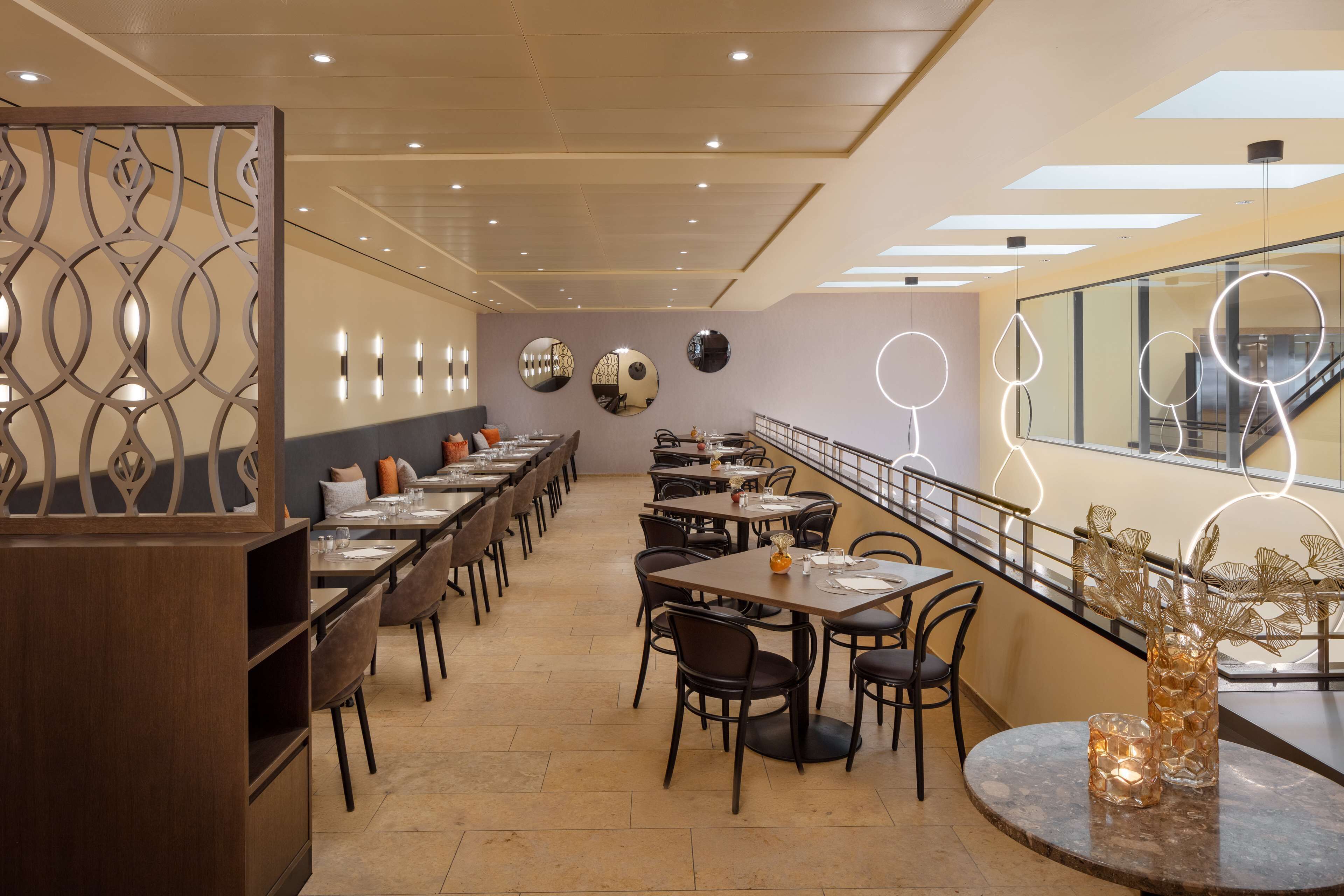A modern restaurant with long tables and chairs, a decorative wall with circular mirrors, and a staircase with glass railings.