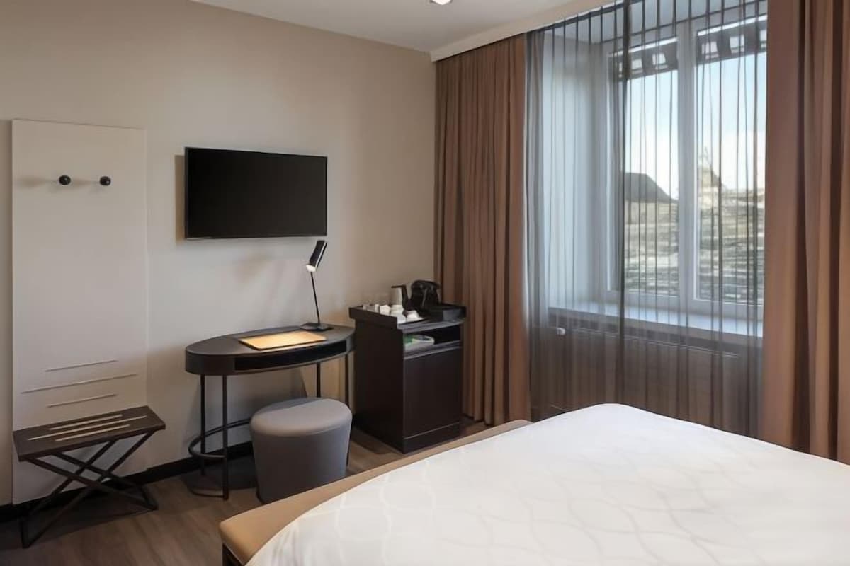 A hotel room with a bed, a desk with a computer, a chair, a small table, a TV, and a window with curtains.