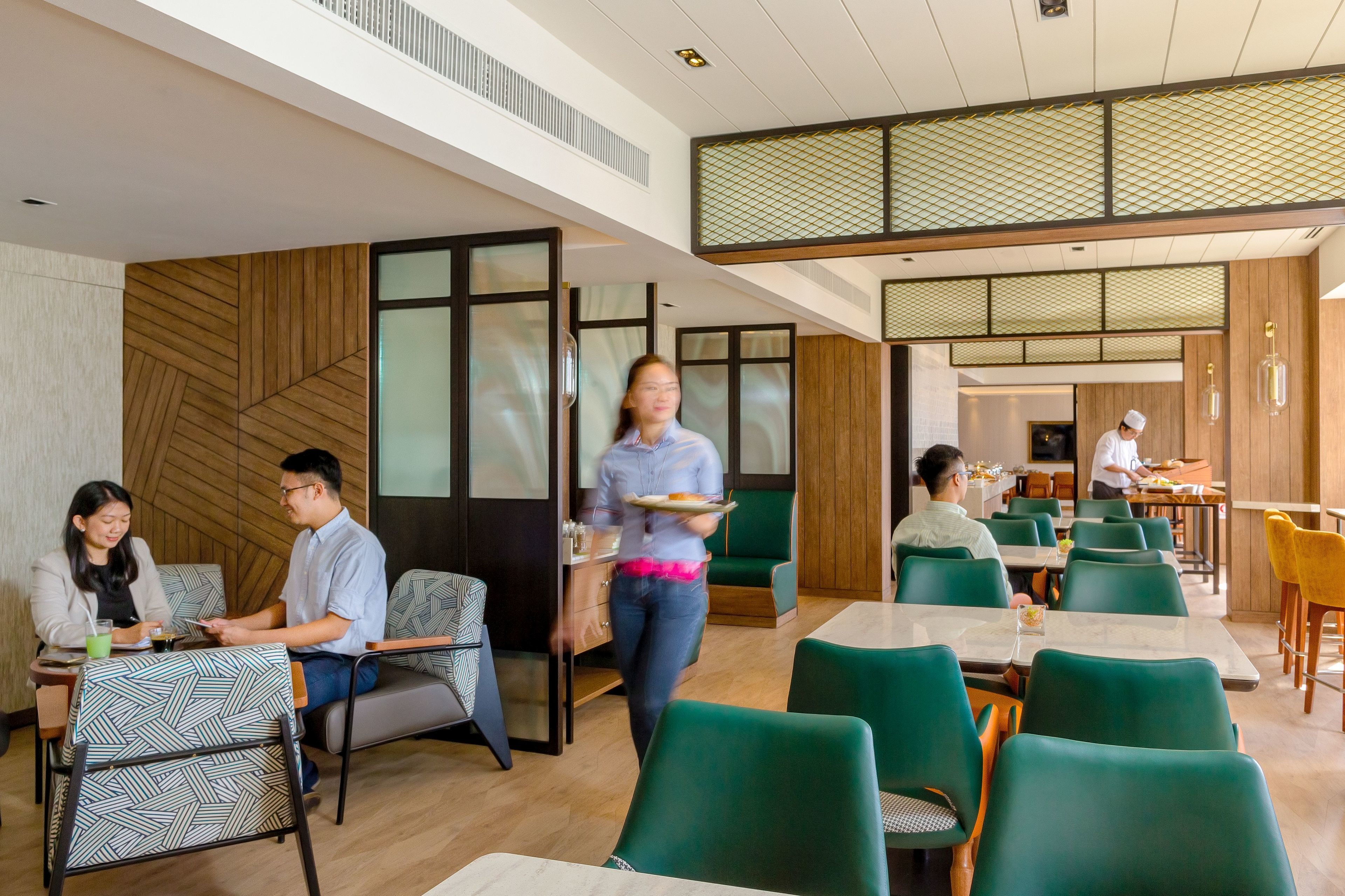 Un espace de restauration moderne avec des chaises et des tables vertes, un serveur portant un plateau, et une personne portant un chapeau de chef qui prépare des plats.