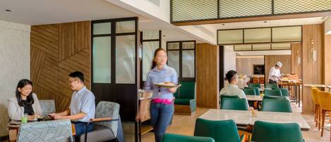 A modern dining area with green chairs and tables, a waiter carrying a tray, and a person in a chef's hat preparing food.
