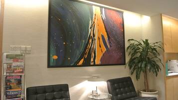 A lobby with two black leather chairs, a round glass table, a large abstract painting, a potted plant, and a wall-mounted information board.