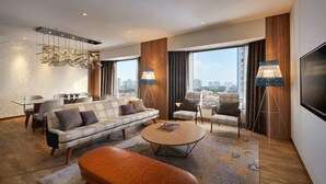 A modern living room with a sofa, armchairs, a coffee table, and a TV. There are large windows with curtains, a city view, and a floor lamp. - JEN Penang Georgetown by Shangri-La (George Town)