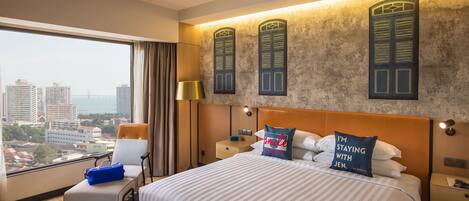 A hotel room with a large bed, a chair, a nightstand, a lamp, and a view of the city through a large window.