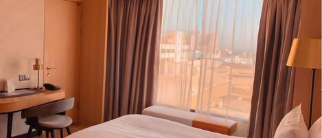 A hotel room with a bed, a desk, and a chair. There is a window with curtains, and a lamp is visible on the right side.