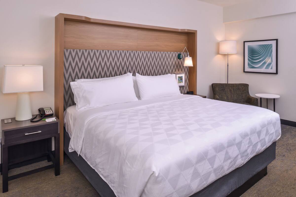 A hotel room with a large bed, a nightstand with a lamp, a chair, and a small table.