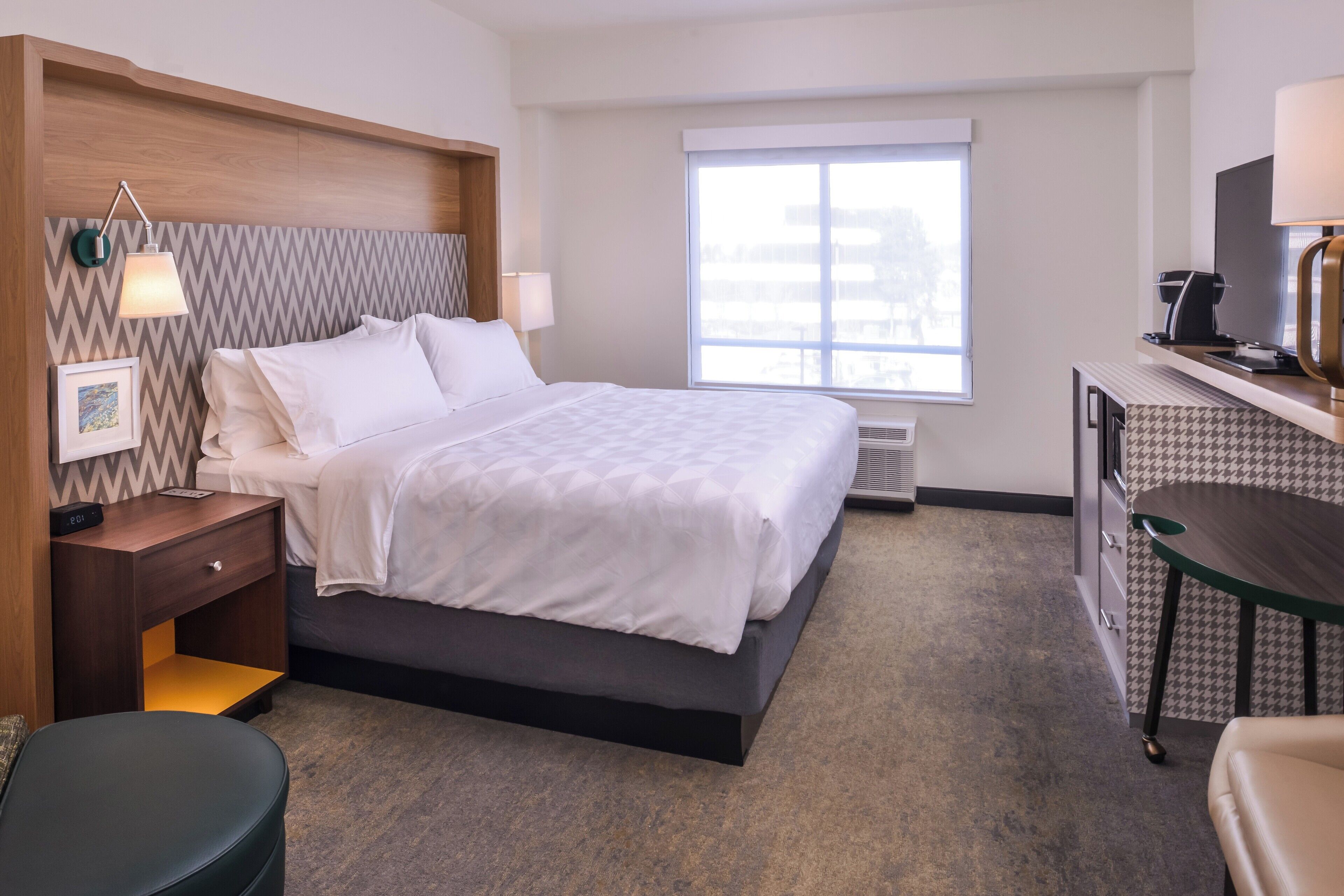 A hotel room with a bed, bedside table, lamp, picture frame, and a desk with a lamp.