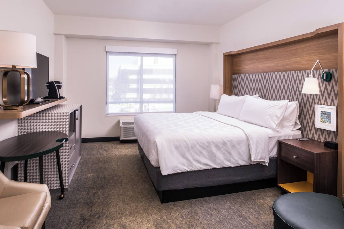 A hotel room with a bed, a desk, a chair, a lamp, and a window.