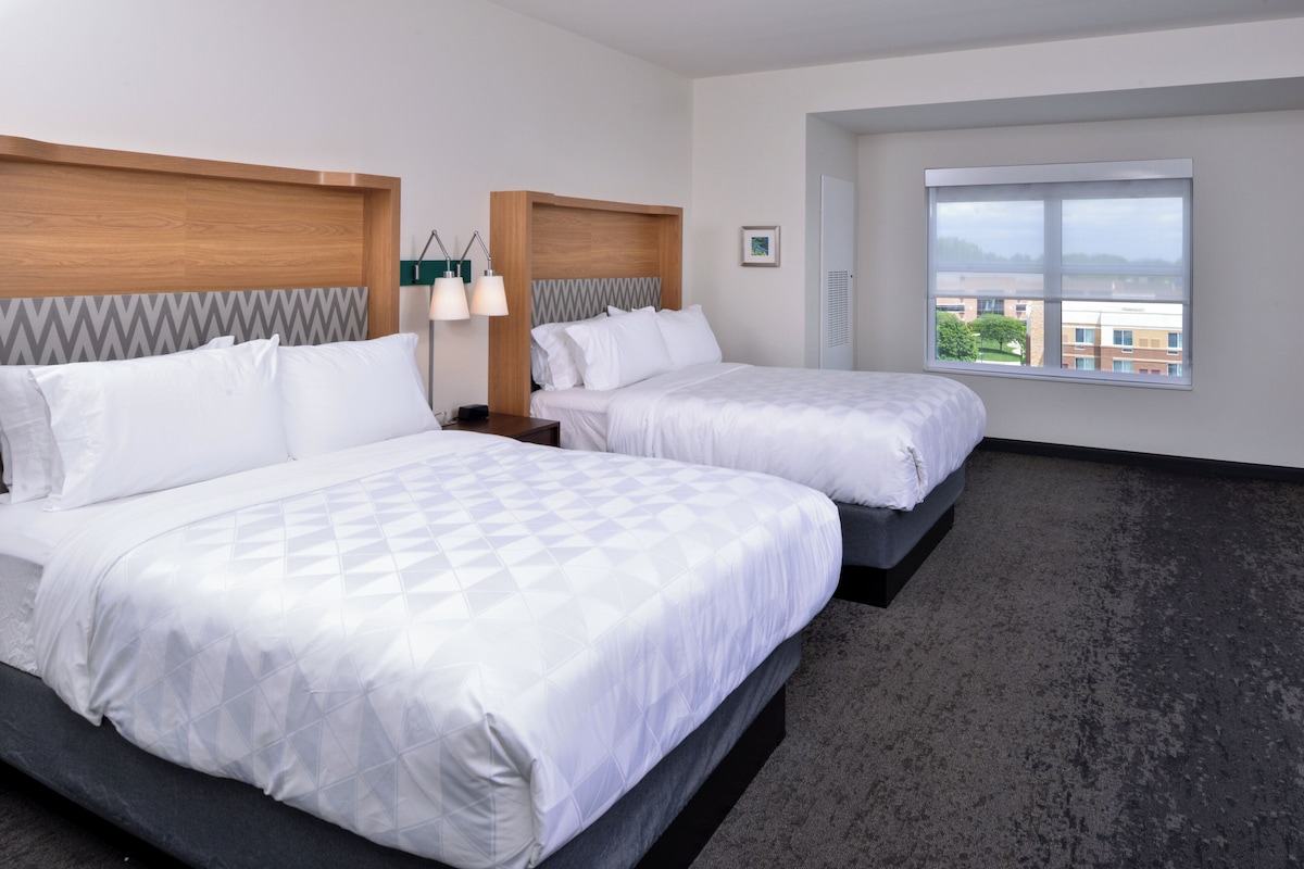 A hotel room with two beds, a large window, and a view of buildings.