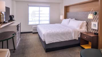 A hotel room with a bed, bedside table, lamp, desk with a coffee maker, and a window.