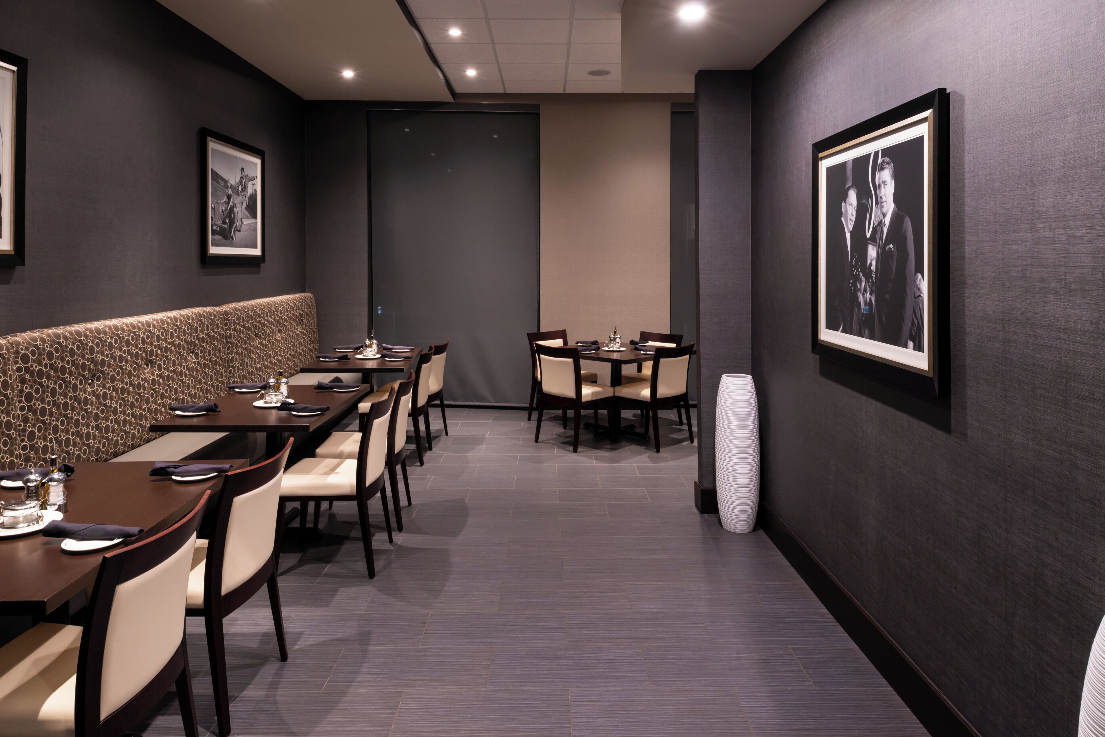 Un restaurant avec un long comptoir, plusieurs tables et des chaises. Les murs sont ornés de tableaux encadrés et un grand vase se trouve sur la droite.