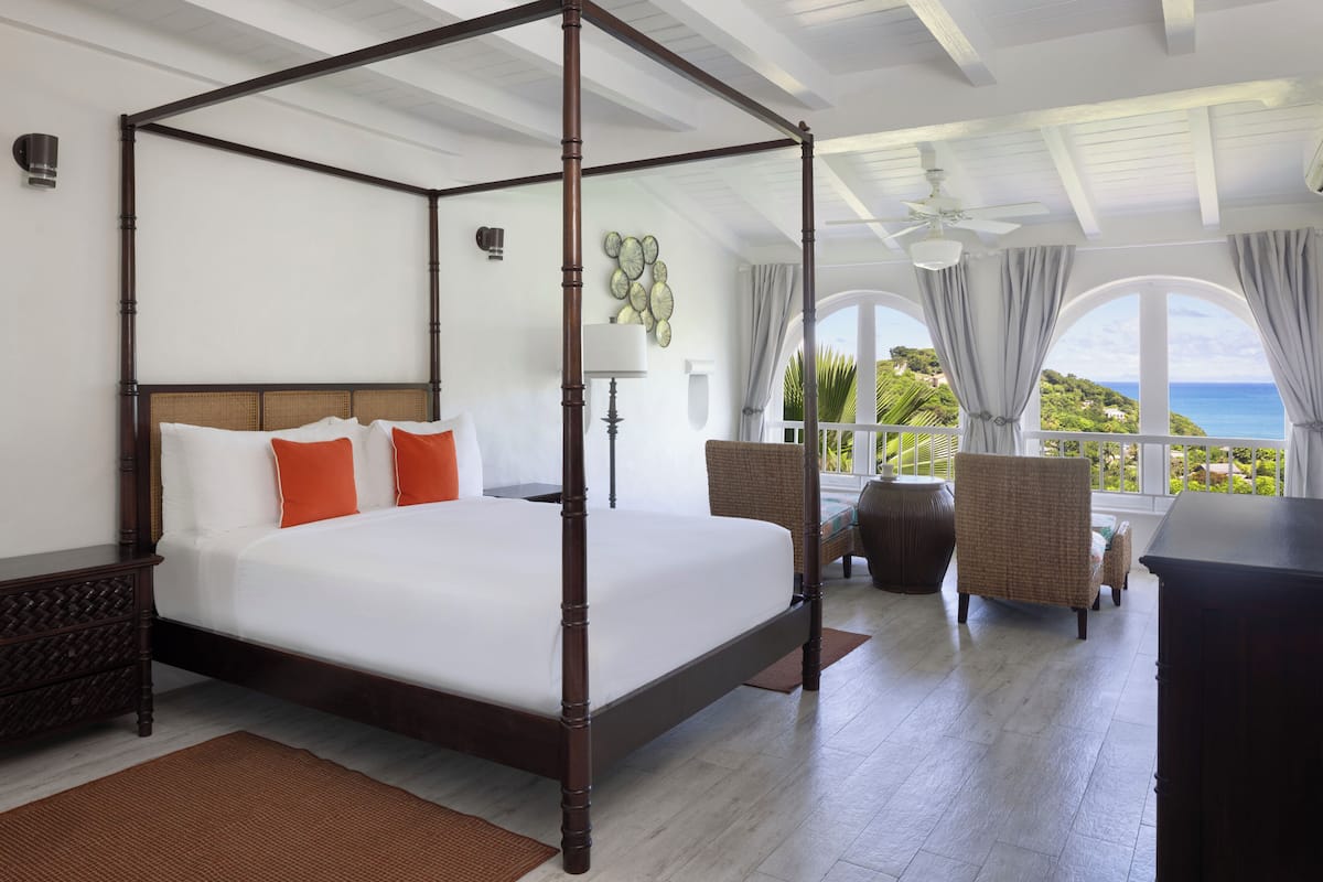 A four-poster bed with white linens and orange pillows, a wicker chair, and a view of the ocean through large windows.