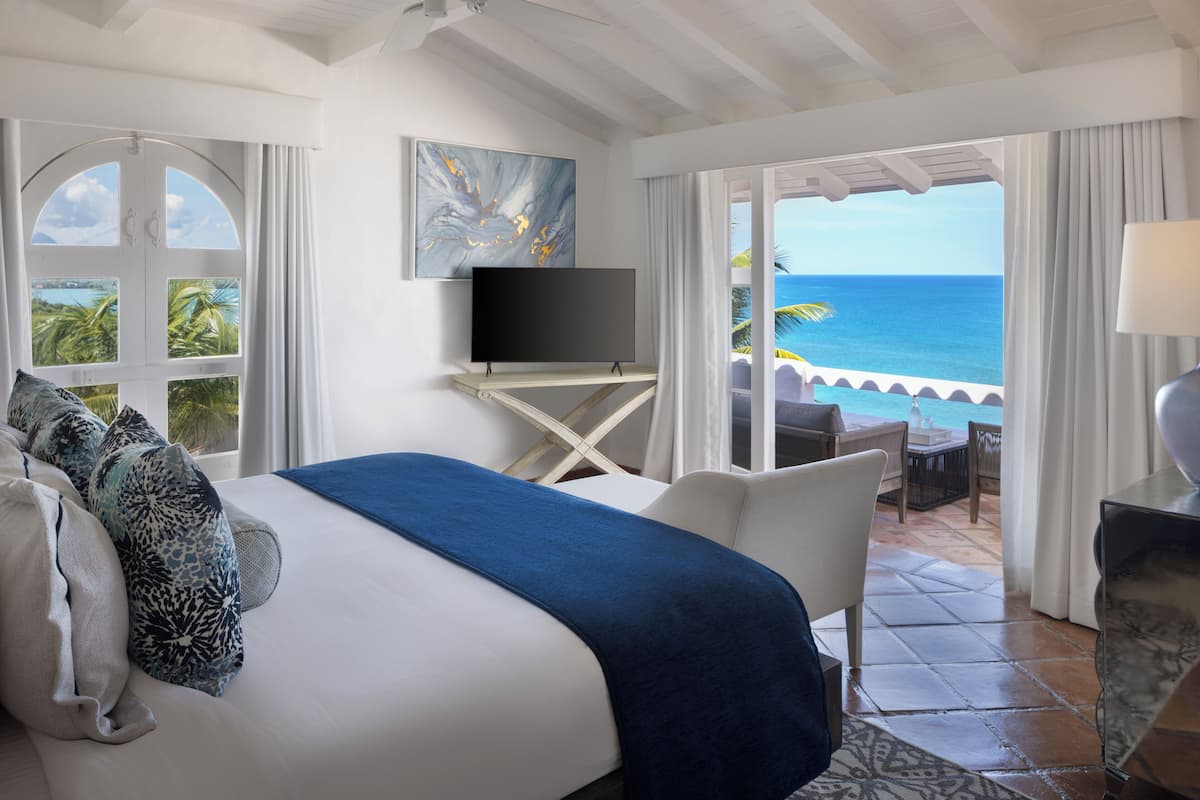 A hotel room with a bed, a TV, a balcony with a view of the ocean, and a seating area with a table and chairs.