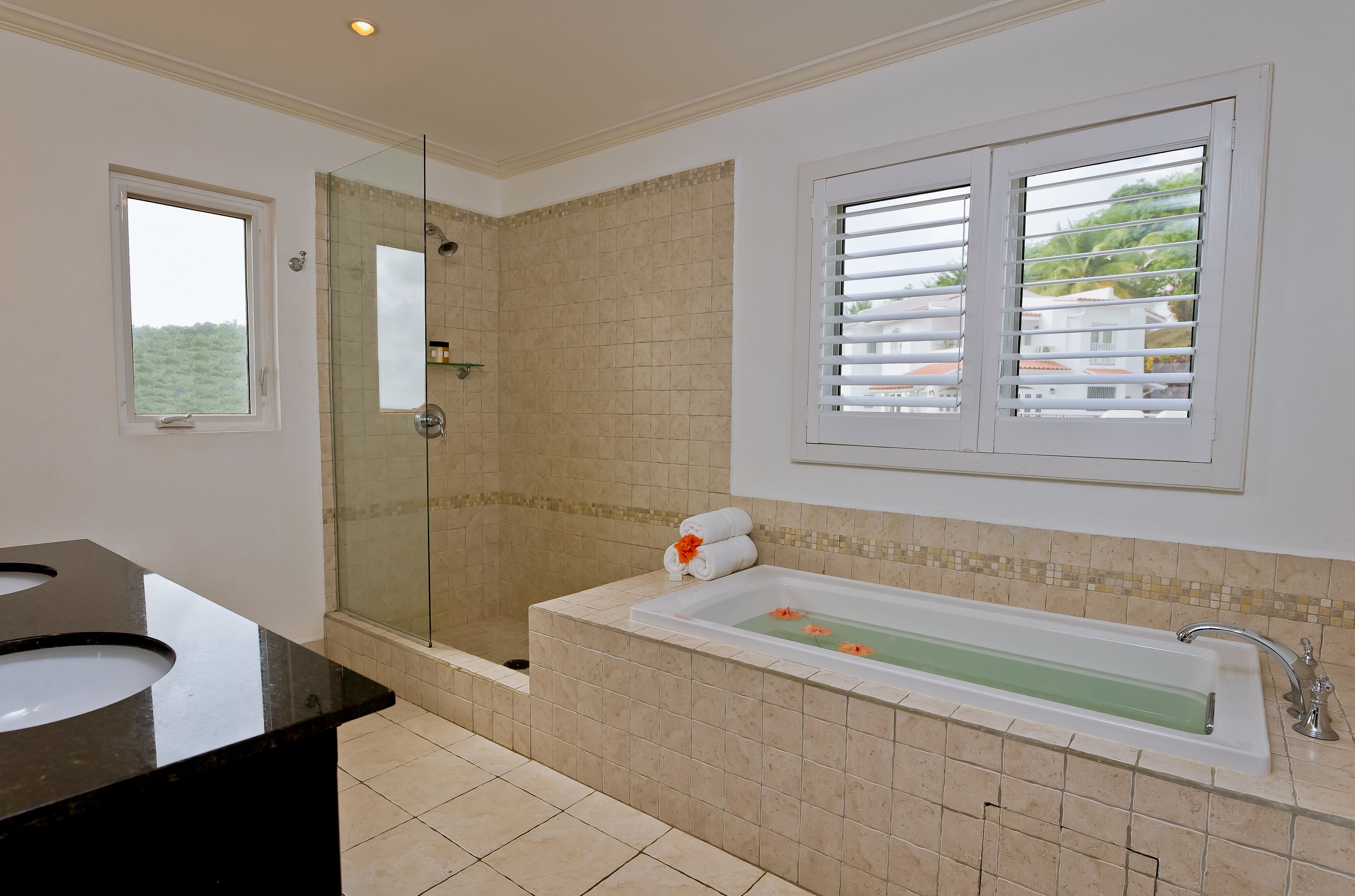 A bathroom with a glass-enclosed shower, a bathtub with a flower arrangement, and a window with blinds.