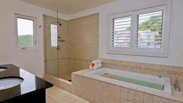 A bathroom with a glass-enclosed shower, a bathtub with a flower arrangement, and a window with blinds.
