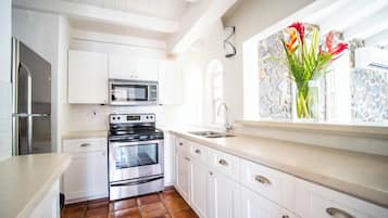 A kitchen with white cabinets, a stainless steel refrigerator, a microwave, an oven, and a window with a stone wall and a vase of flowers.