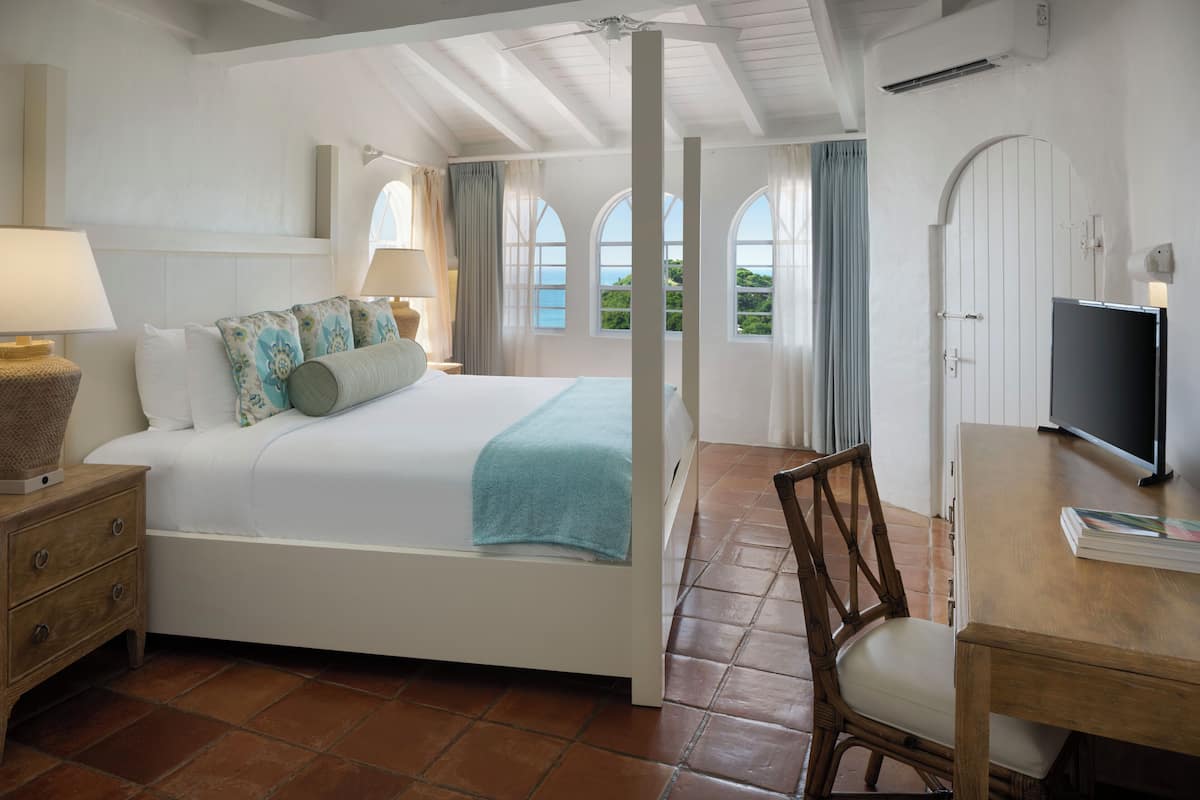 A bedroom with a bed, bedside table, chair, TV, and windows with curtains.