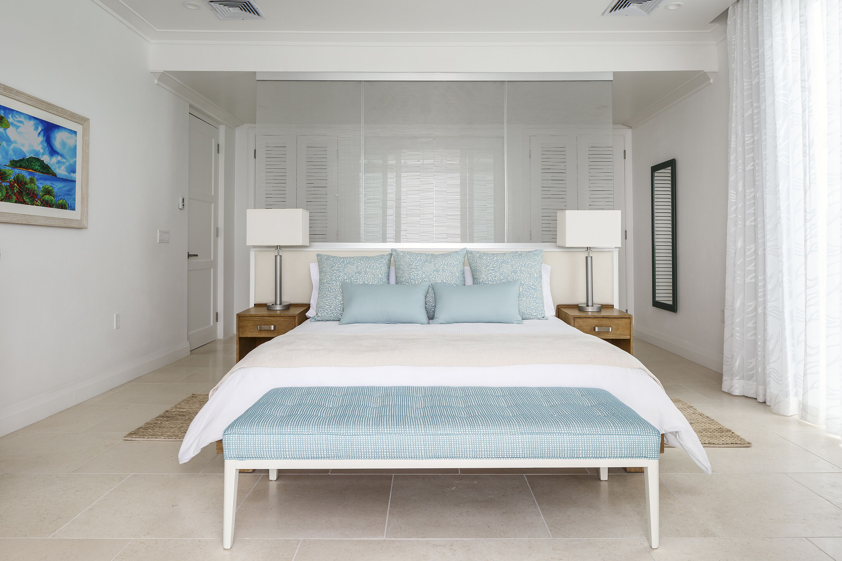 Een slaapkamer met een groot bed, twee nachtkastjes, een bankje, een schilderij en een raam met jaloezieën.
