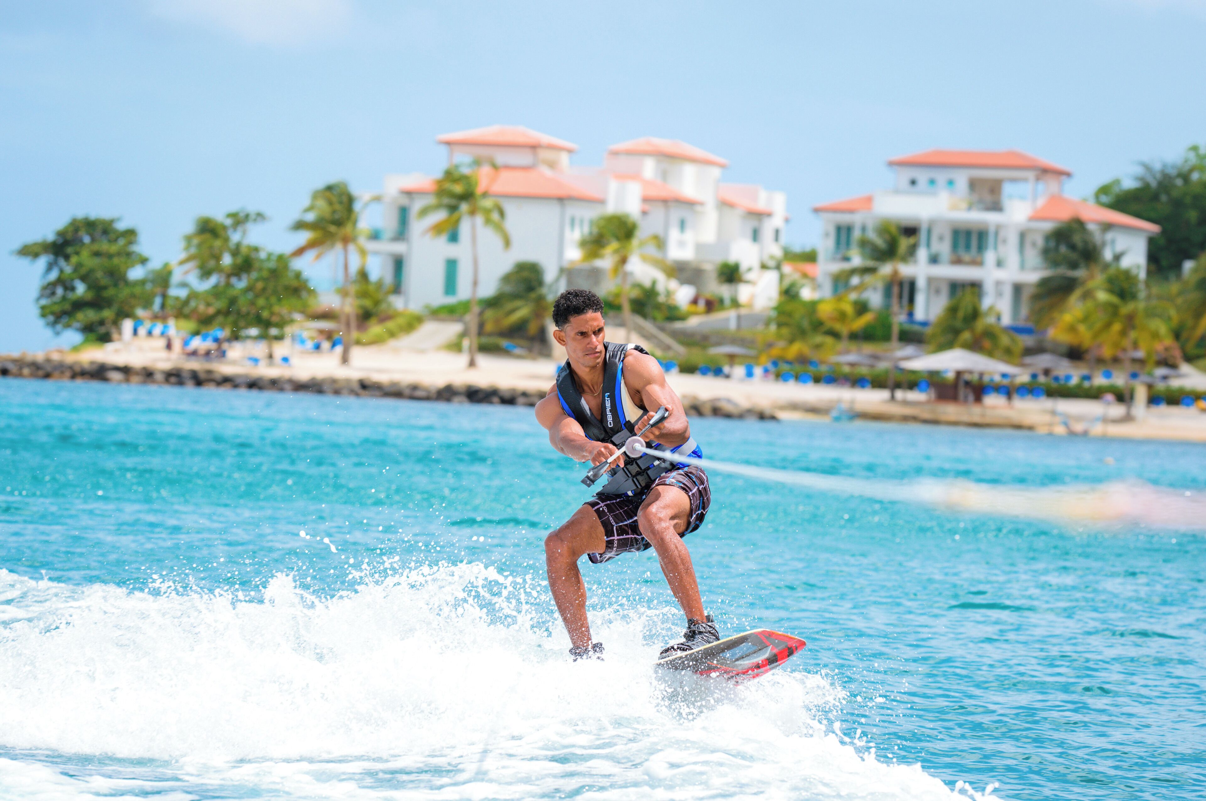 Una persona practicando wakeboard en un día despejado, con una playa y edificios de un complejo turístico al fondo.